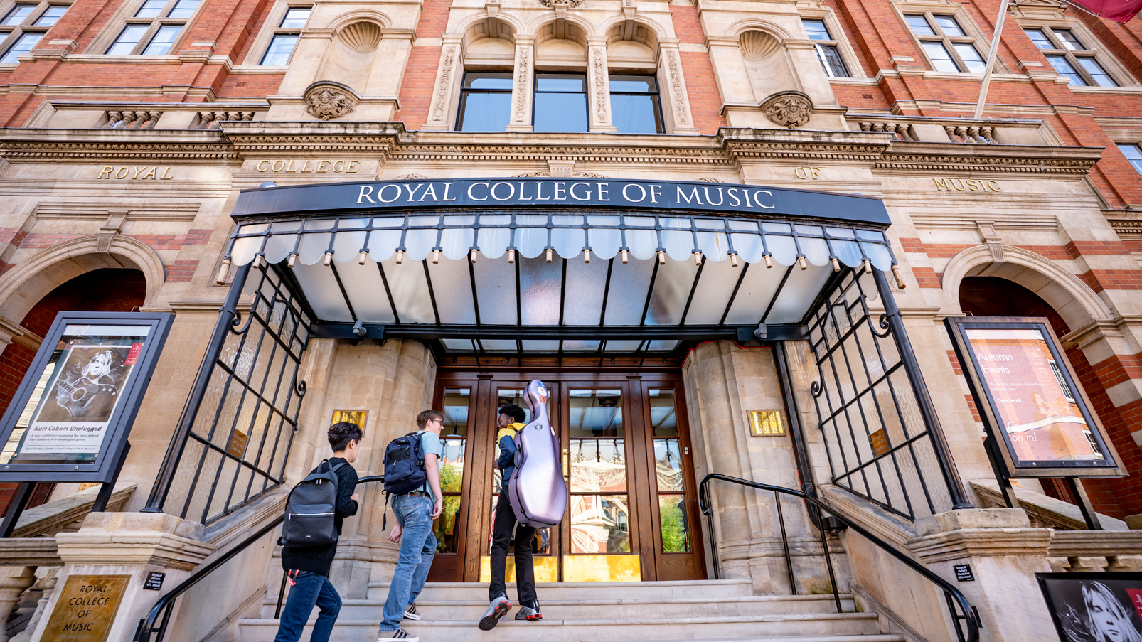 A group of students wearing casual clothes, with students holding instrument cases walking into the building of the Royal College of Music, an old, red brick, Victorian building, in the background.